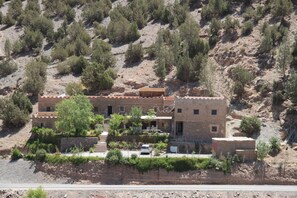 Un edificio de dos pisos con techo plano, rodeado de vegetación escasa y terreno rocoso.