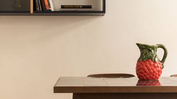 A wooden table with a red fruit-shaped pitcher on it, a bookshelf with books and a decorative item above, and a plain wall in the background.