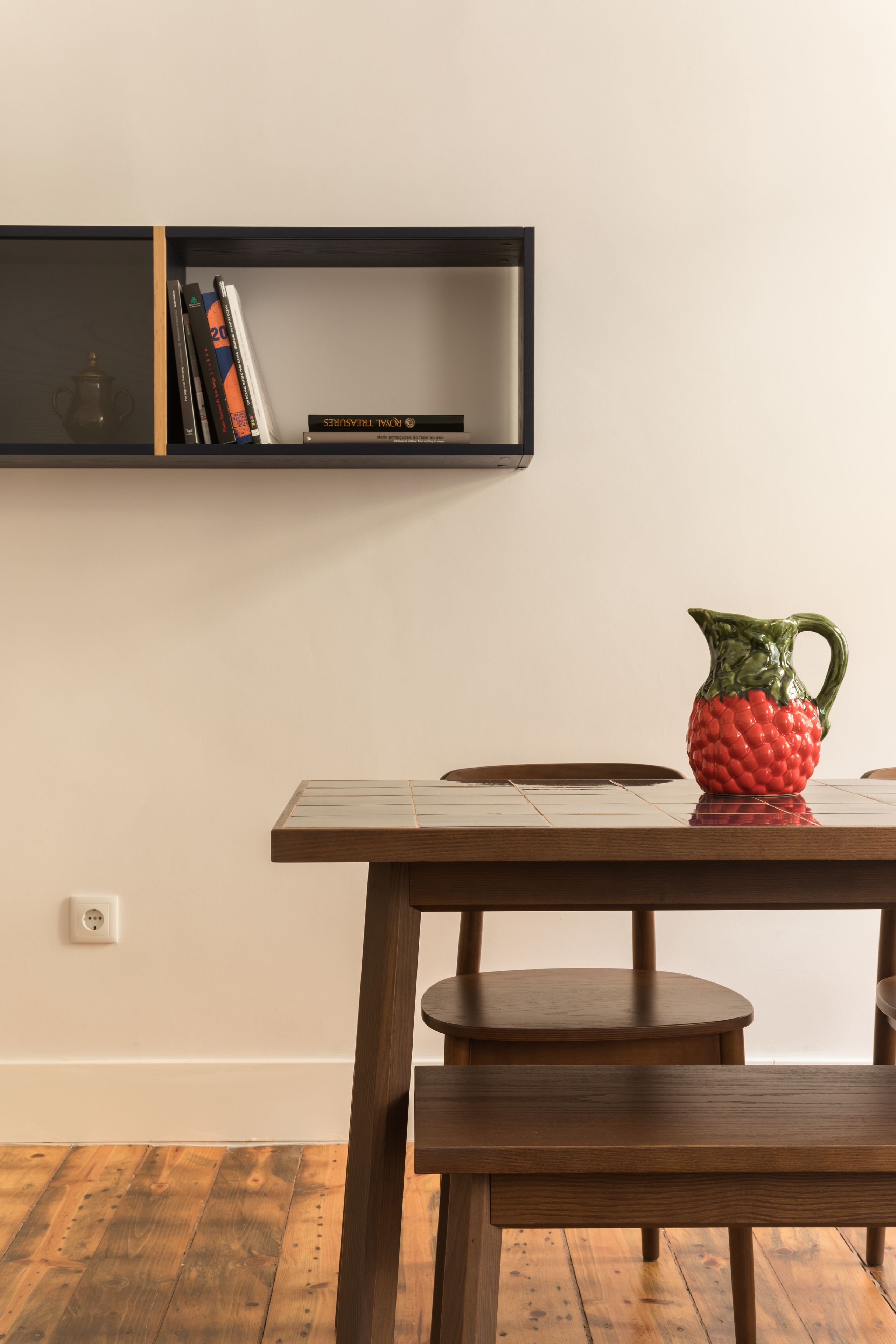Une table en bois sur laquelle se trouve un pichet rouge en forme de fruit, une bibliothèque remplie de livres et un petit tabouret rond.