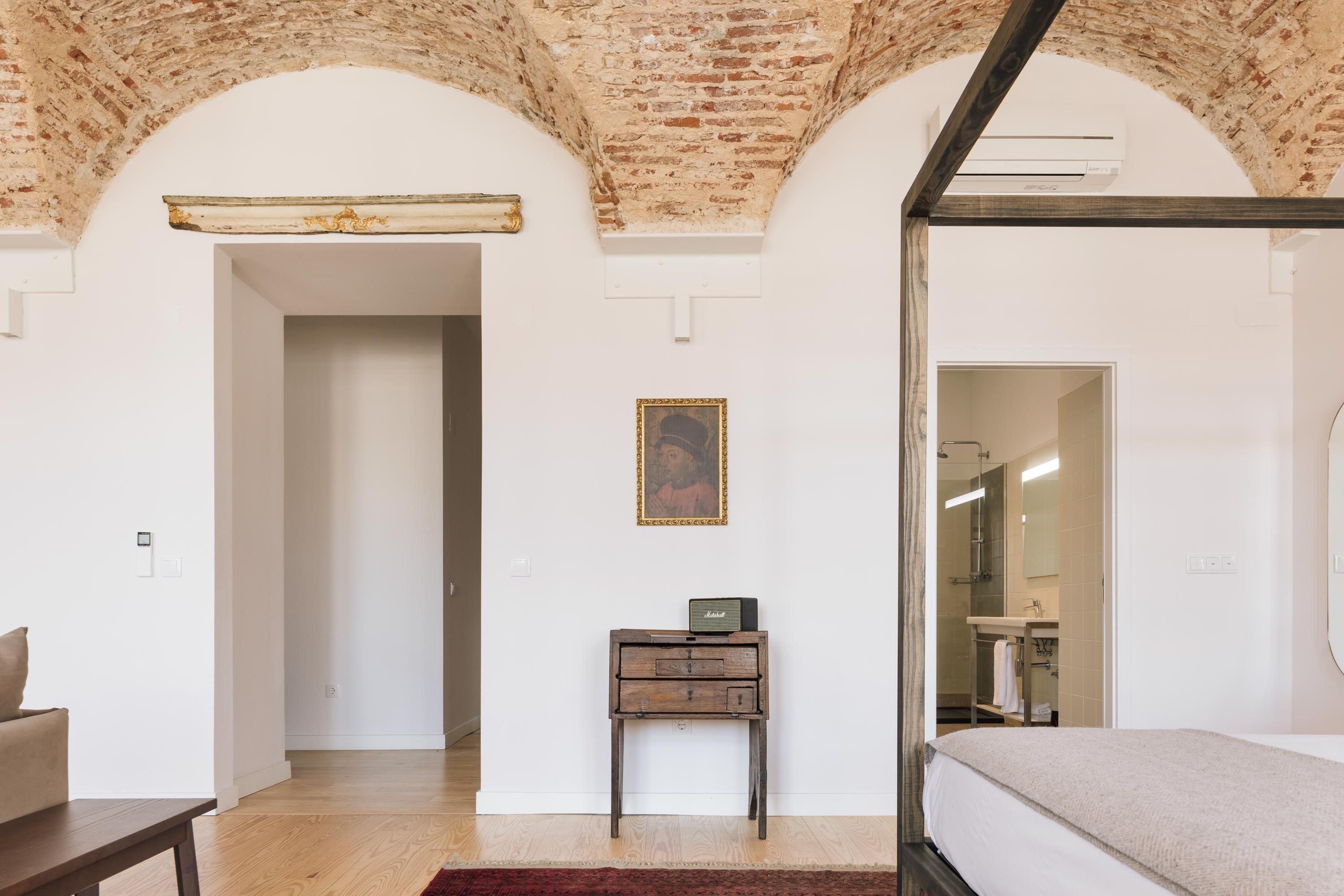 A bedroom with a brick ceiling, a bed, a small wooden nightstand with a lamp, and a bathroom visible through an open door.