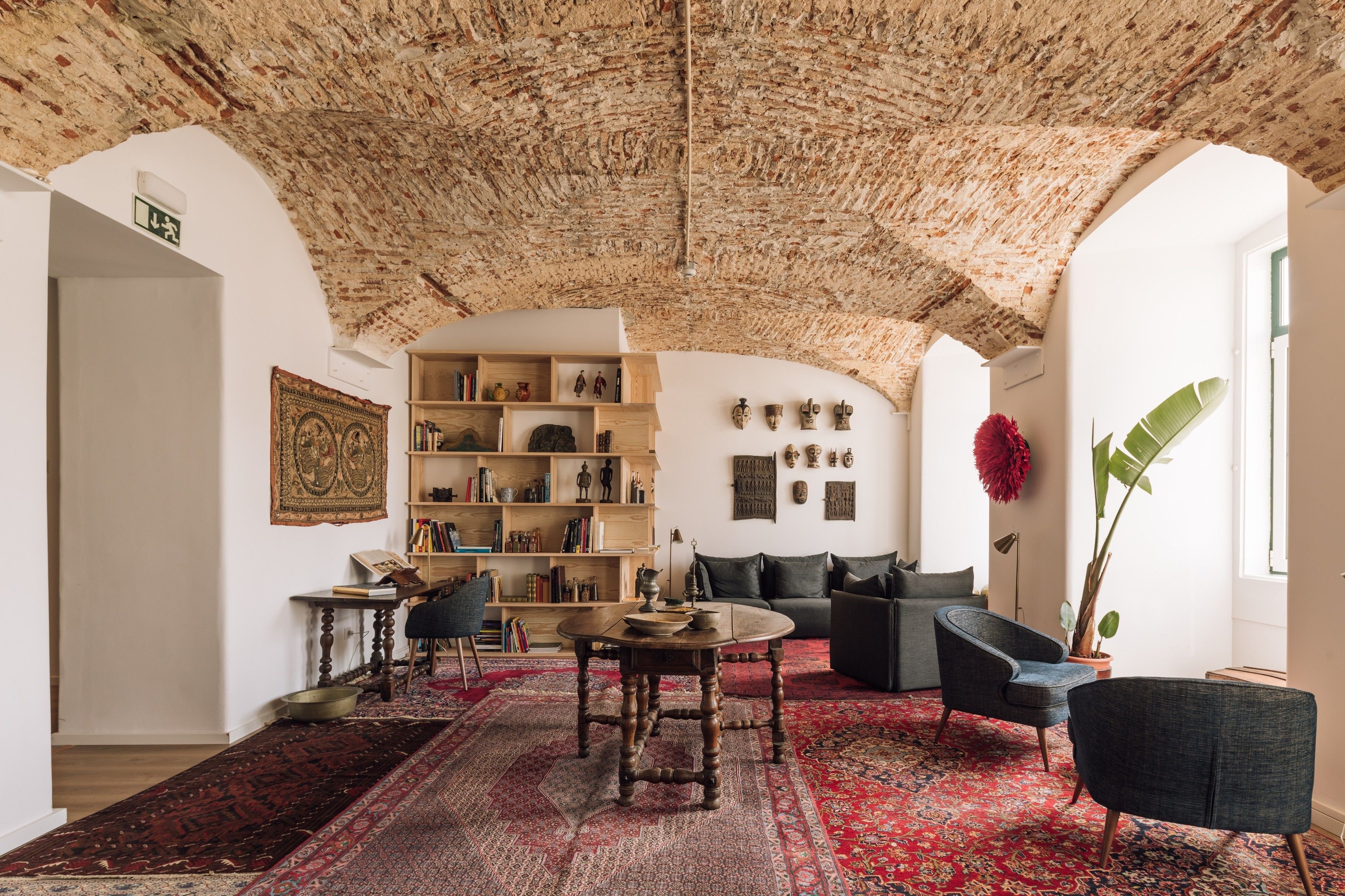 Een ruime woonkamer met een hoog, gestructureerd plafond, een groot rood tapijt met dessin, een houten salontafel, een donkere bank, een boekenkast vol boeken en decoratieve objecten, en een potplant.