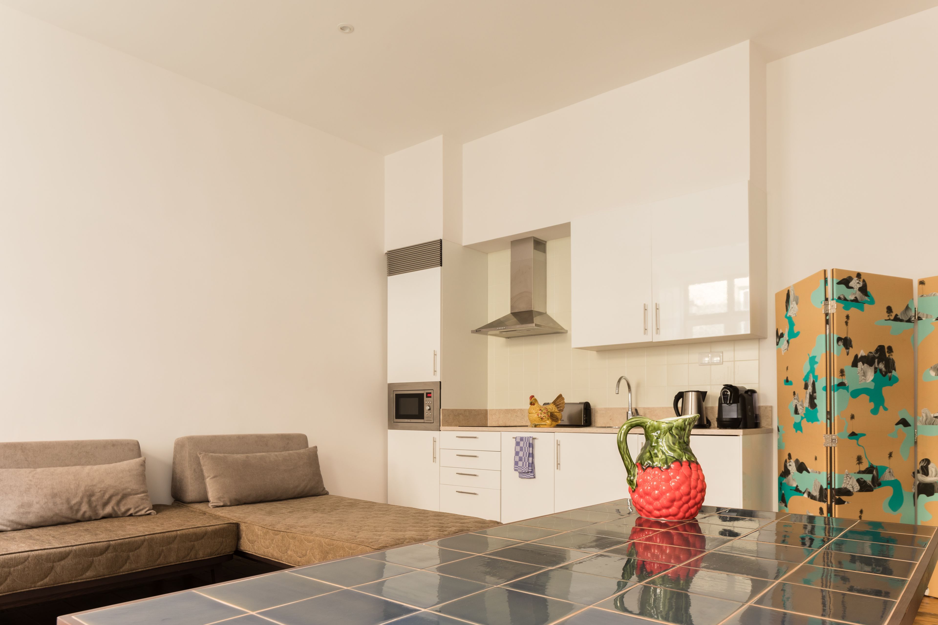 Una cocina moderna con armarios blancos, microondas y cafetera. Hay un sofá con cojines y piso de baldosas.