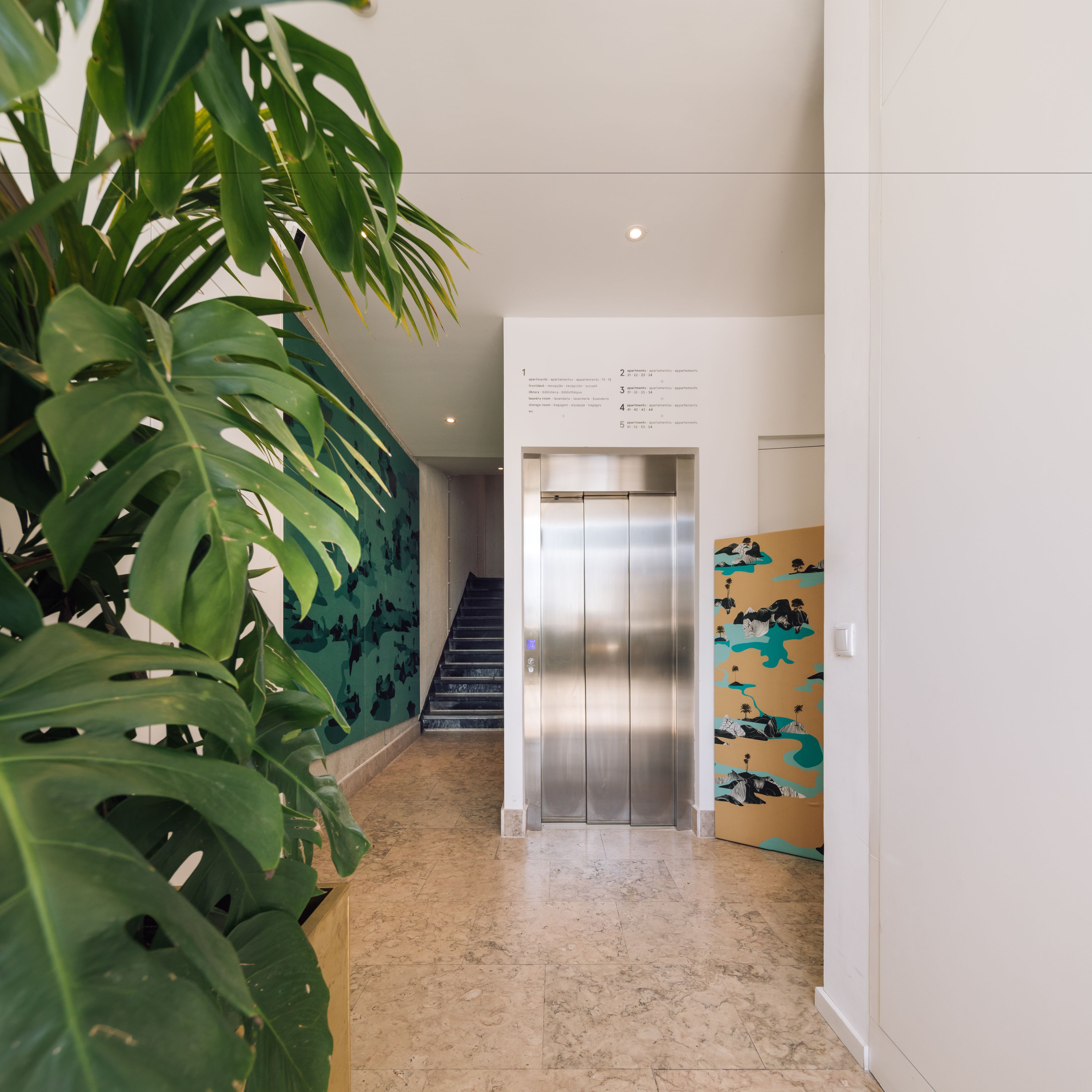 A hallway with a staircase, a metallic elevator, and a wall with a patterned mural.