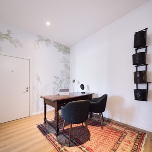 Ein Zimmer mit einem Holztisch, zwei Stühlen, einem Laptop, einer Wand mit Dekoration und einem gemusterten Teppich.