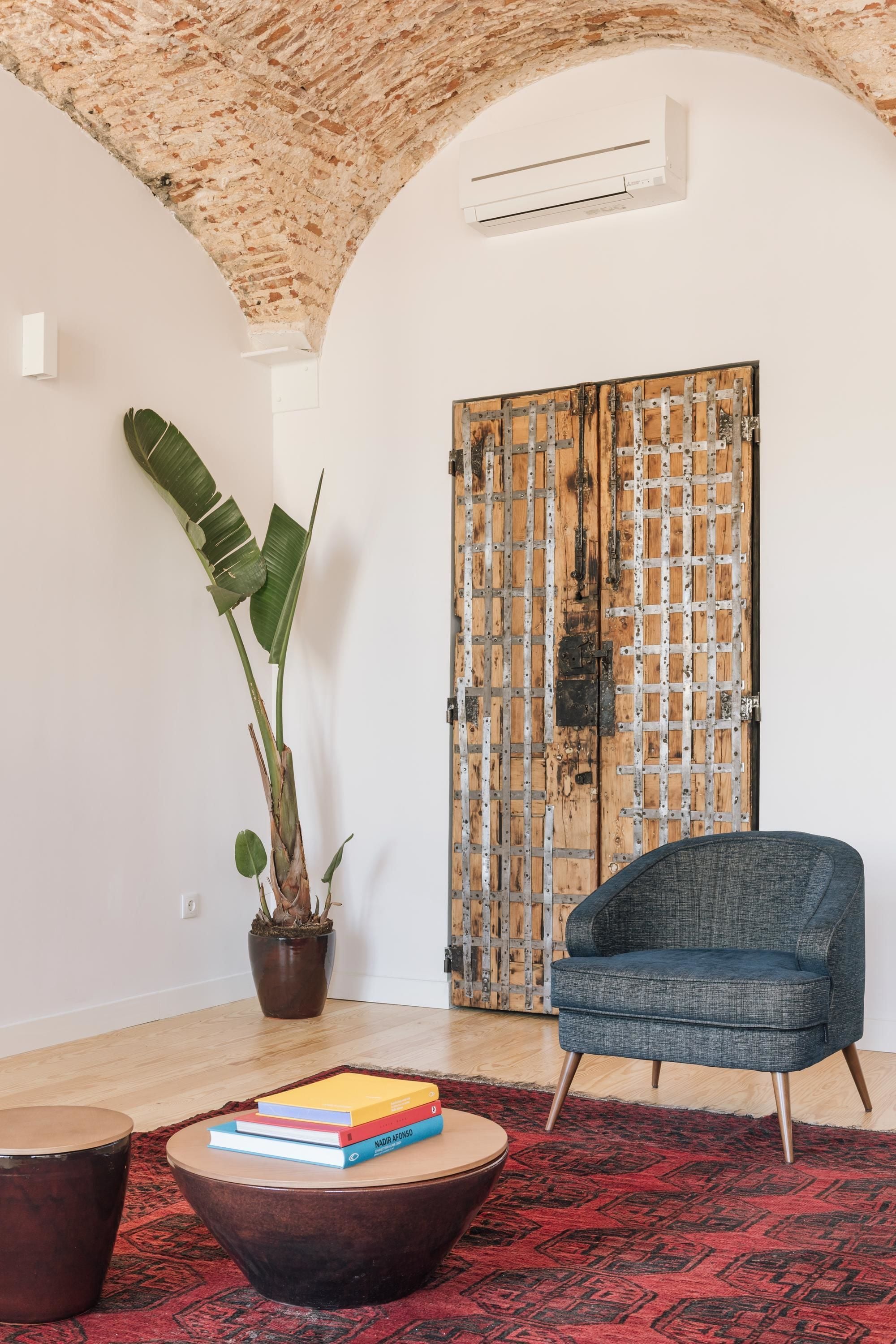 Una stanza con soffitto in mattoni rustici, una porta in legno rinforzata con elementi metallici, una pianta in vaso, una poltrona blu e un tavolino rotondo con libri.
