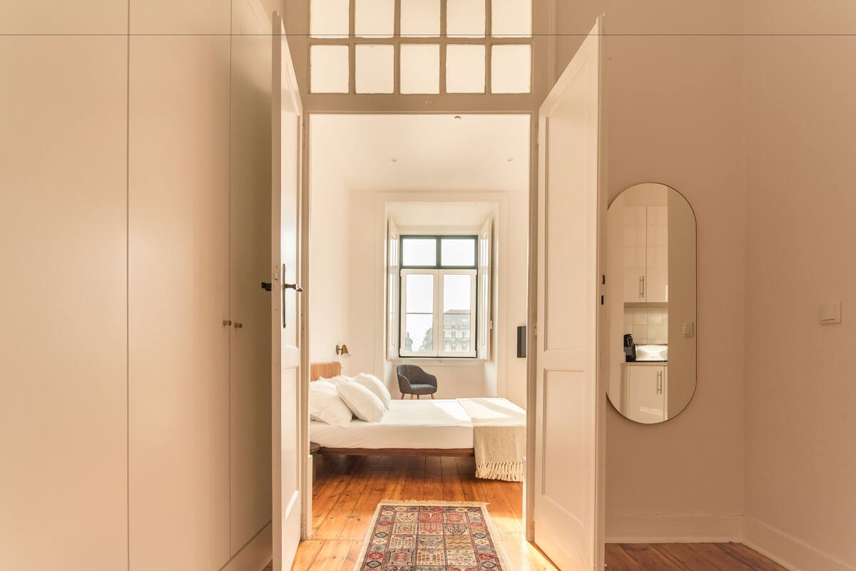 Un dormitorio con una cama, una silla, un ventanal y un espejo, con vistas a una cocina.