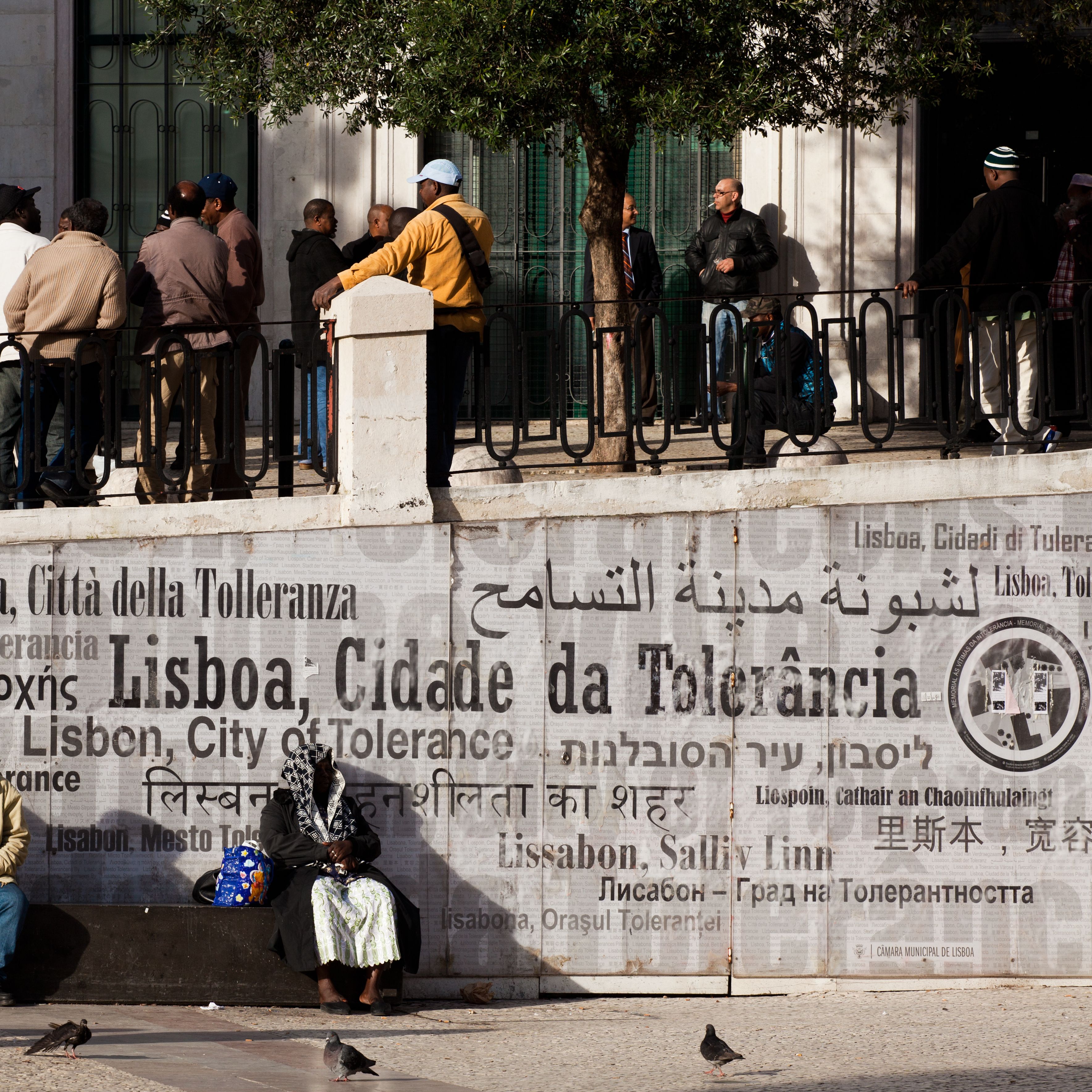 A group of people standing and sitting near a wall with a sign in multiple languages.