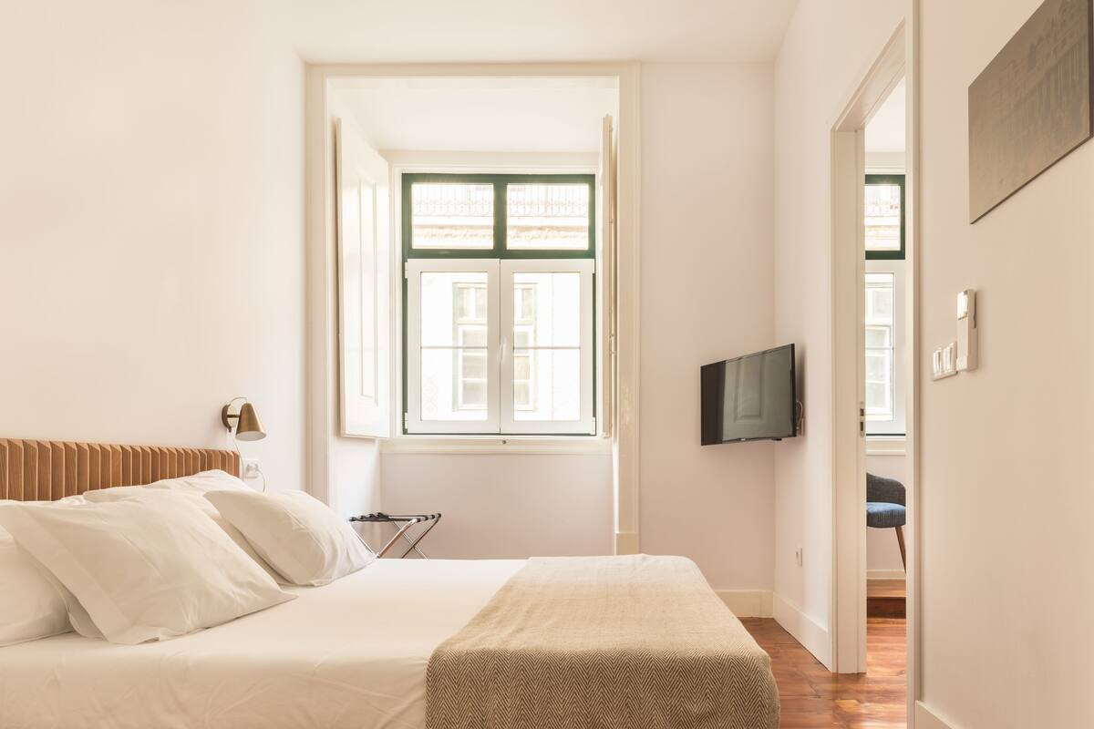 Un dormitorio con cama, televisión, ventana y puerta que da a otra habitación.