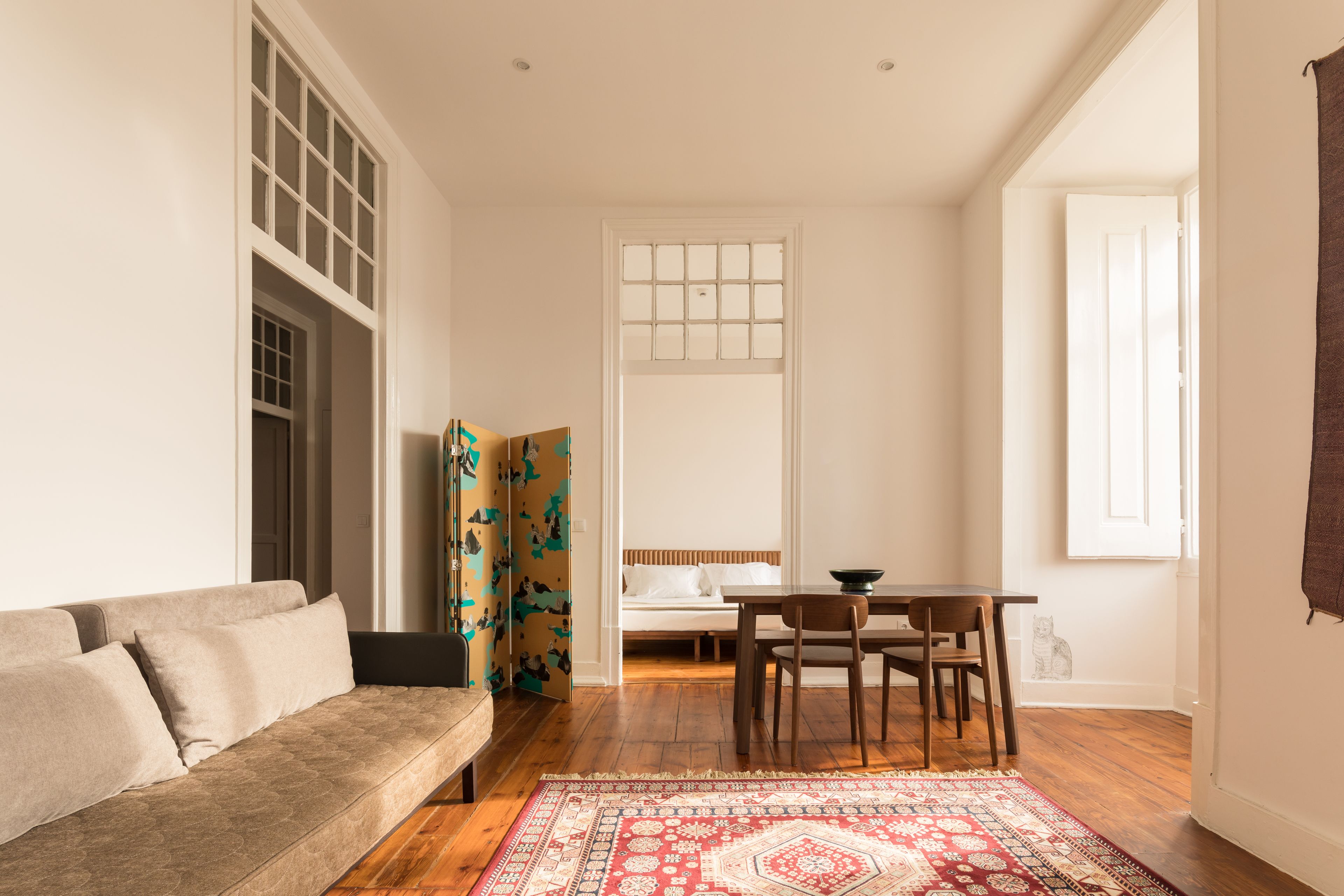 Ein Wohnzimmer mit einer Couch, einem Esstisch mit Stühlen, einem gemusterten Teppich und einem Fenster mit Vorhängen.