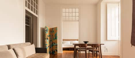 A living room with a sofa, a dining table with chairs, a patterned rug, and a window with curtains.