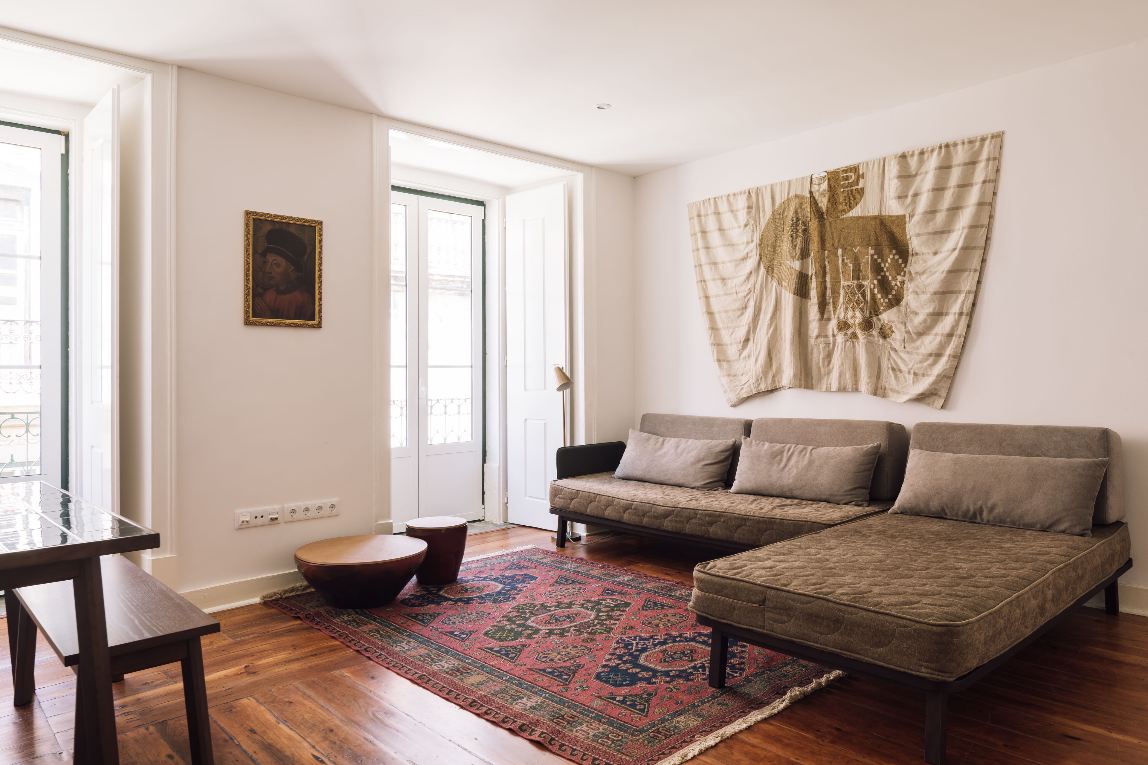 A living room with a sofa, ottoman, and a patterned rug. There is a painting on the wall and a door leading outside.