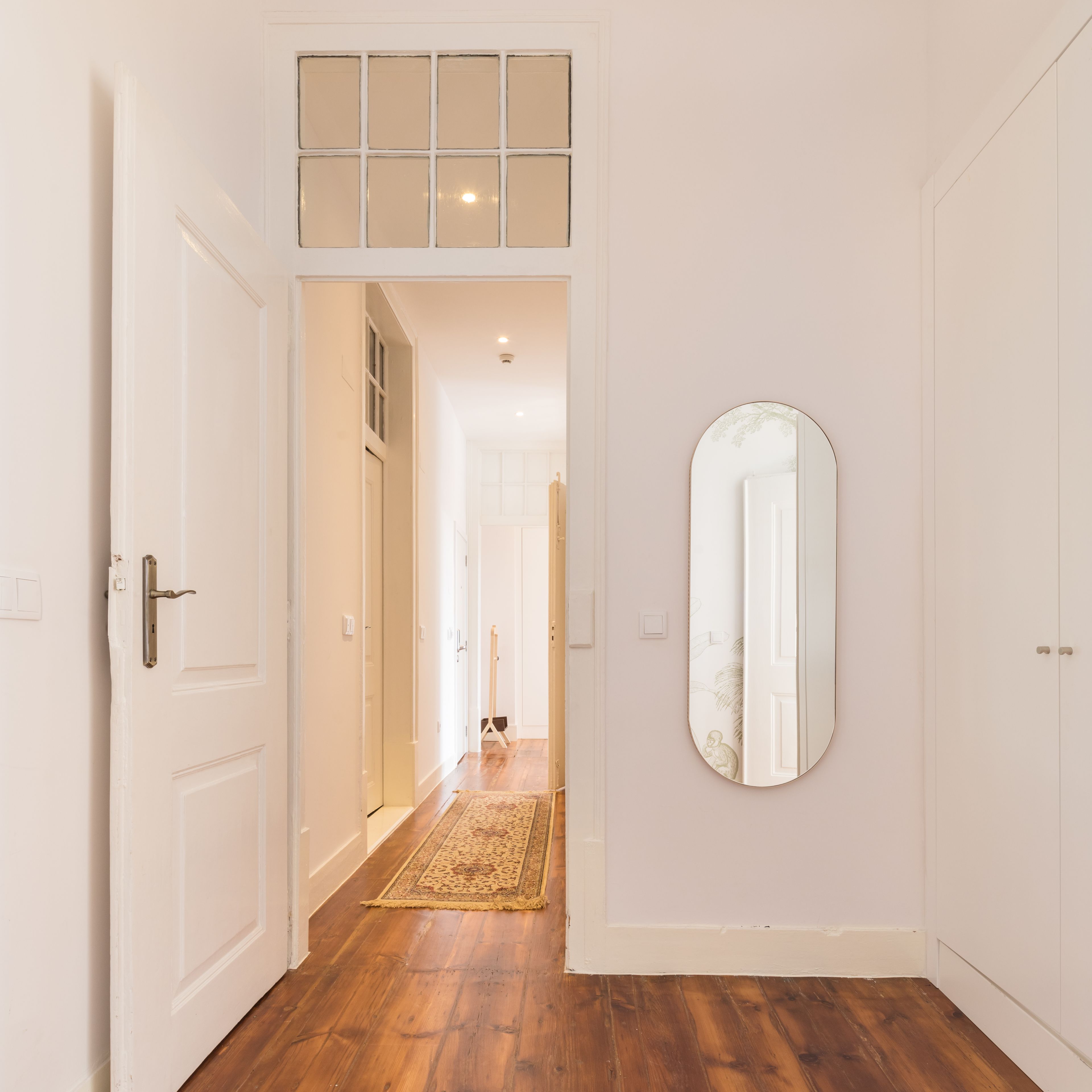A hallway with a wooden floor, white walls, a large mirror, and a door leading to another room.