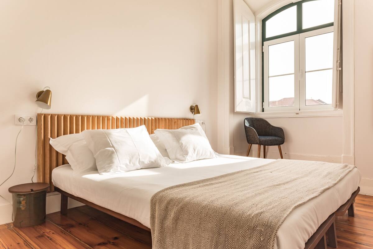 Un dormitorio con suelo de madera, una cama con ropa de cama blanca, una manta estampada en patrón de espiga, una silla y una ventana con vistas.