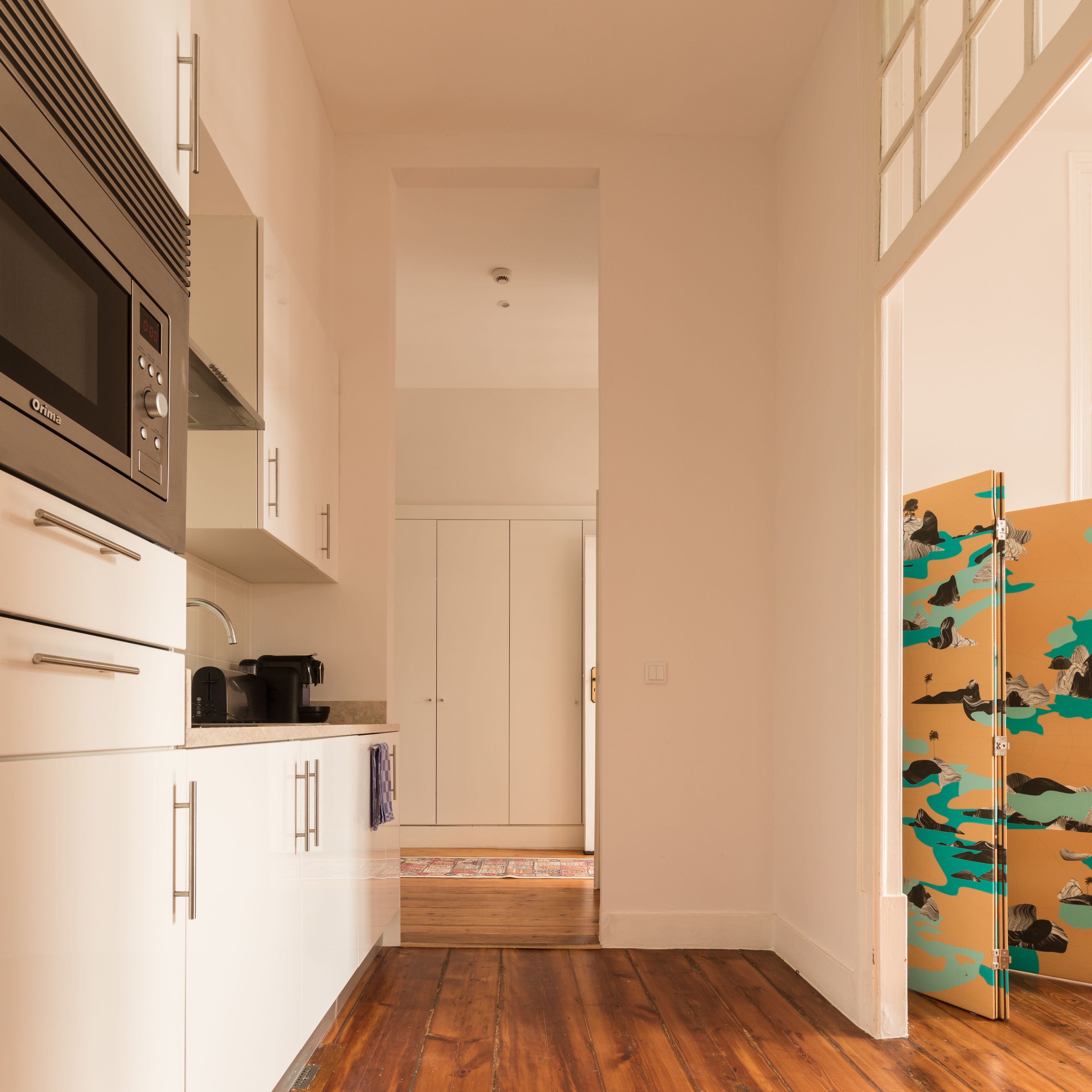 A narrow hallway with a kitchen area on one side, featuring a microwave, cabinets, and a coffee maker. A wooden floor and a door with a colorful mural are visible.