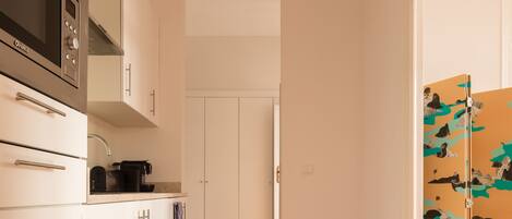 A narrow hallway with a kitchen area on one side, featuring a microwave, cabinets, and a coffee maker. A wooden floor and a door with a colorful mural are visible.