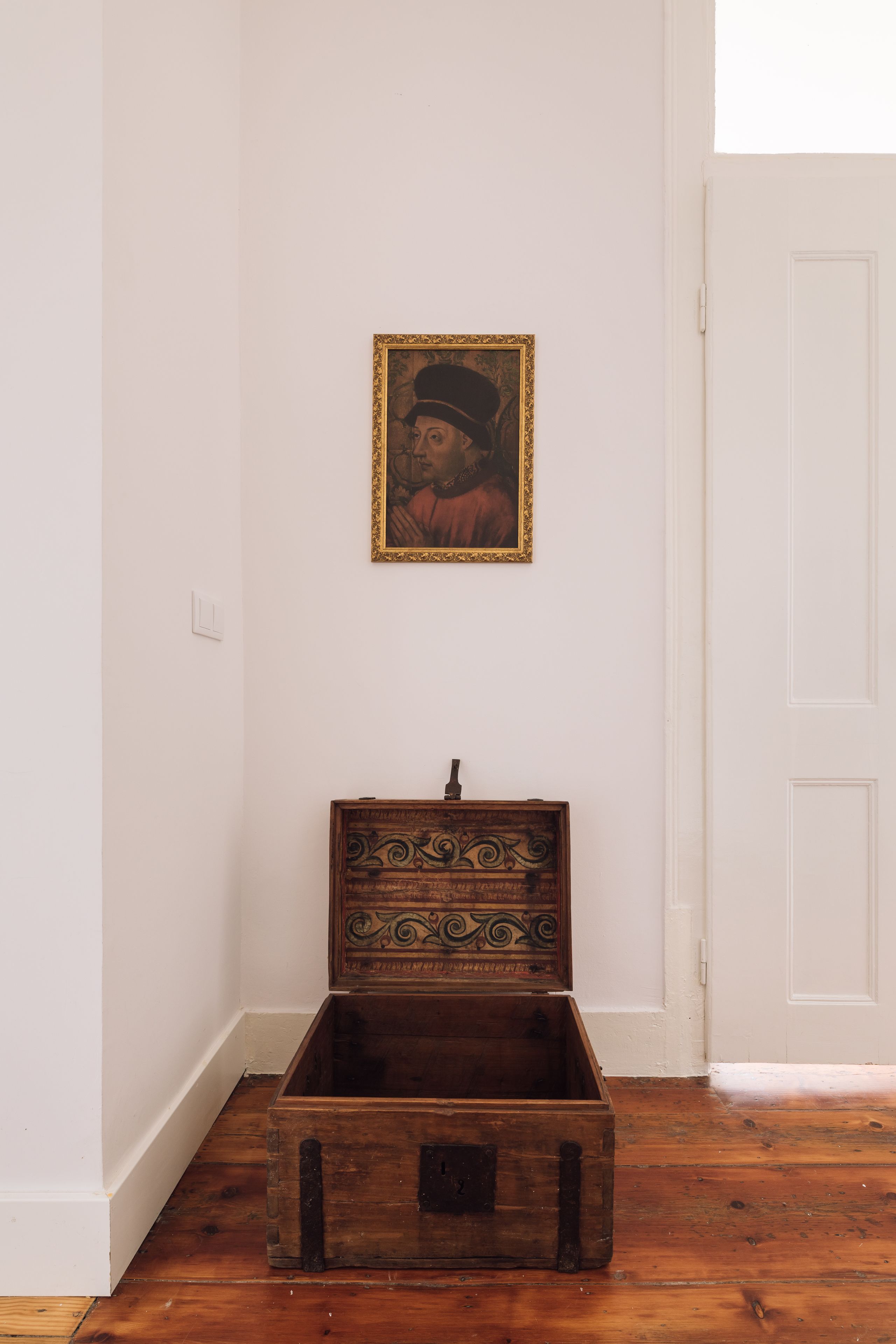 A wooden chest with intricate carvings against a white wall, positioned in a corner with a white door nearby.