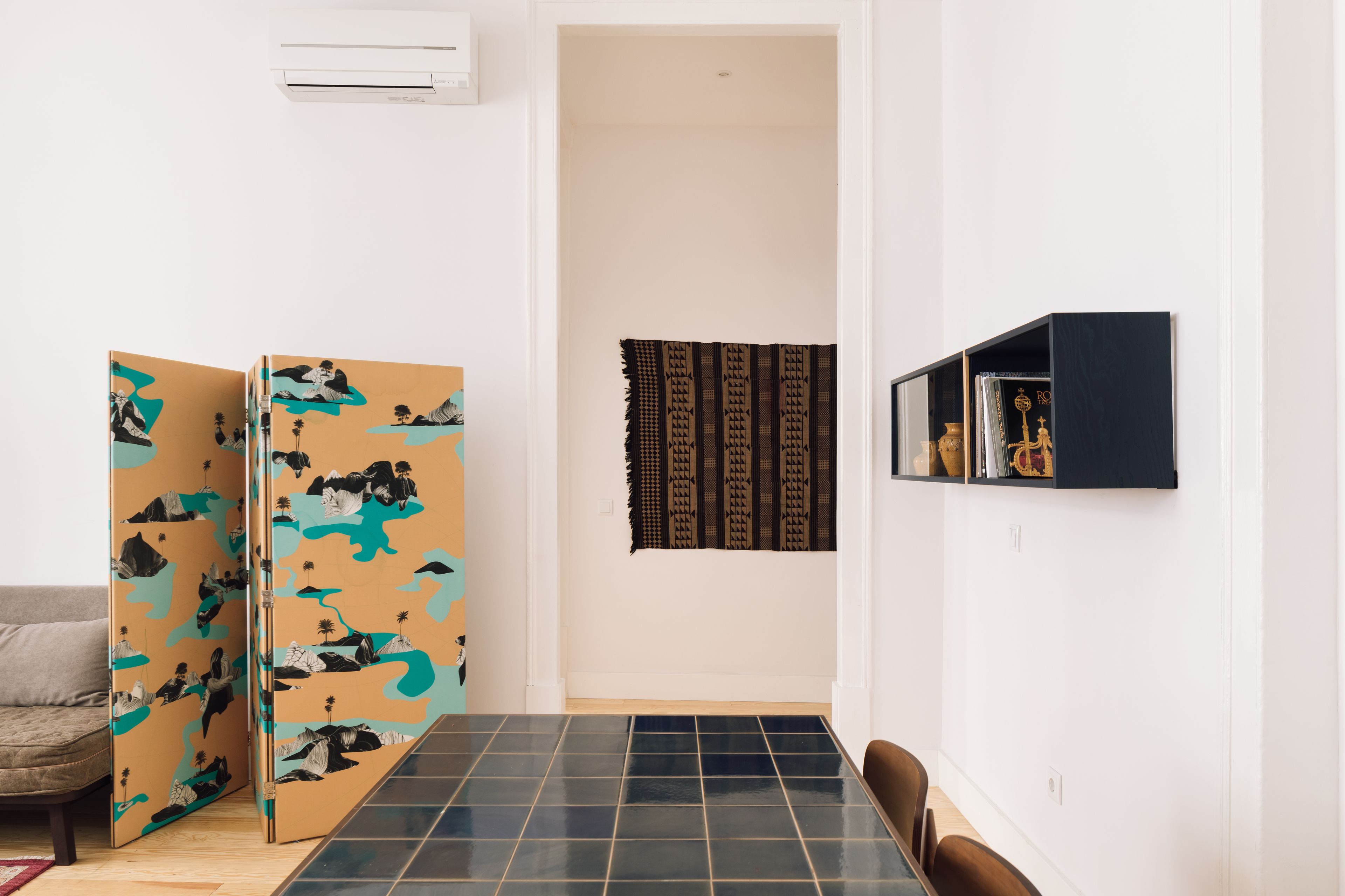 A room with a tiled floor, a sofa, a dining table, a wall-mounted shelf, and a patterned tapestry.