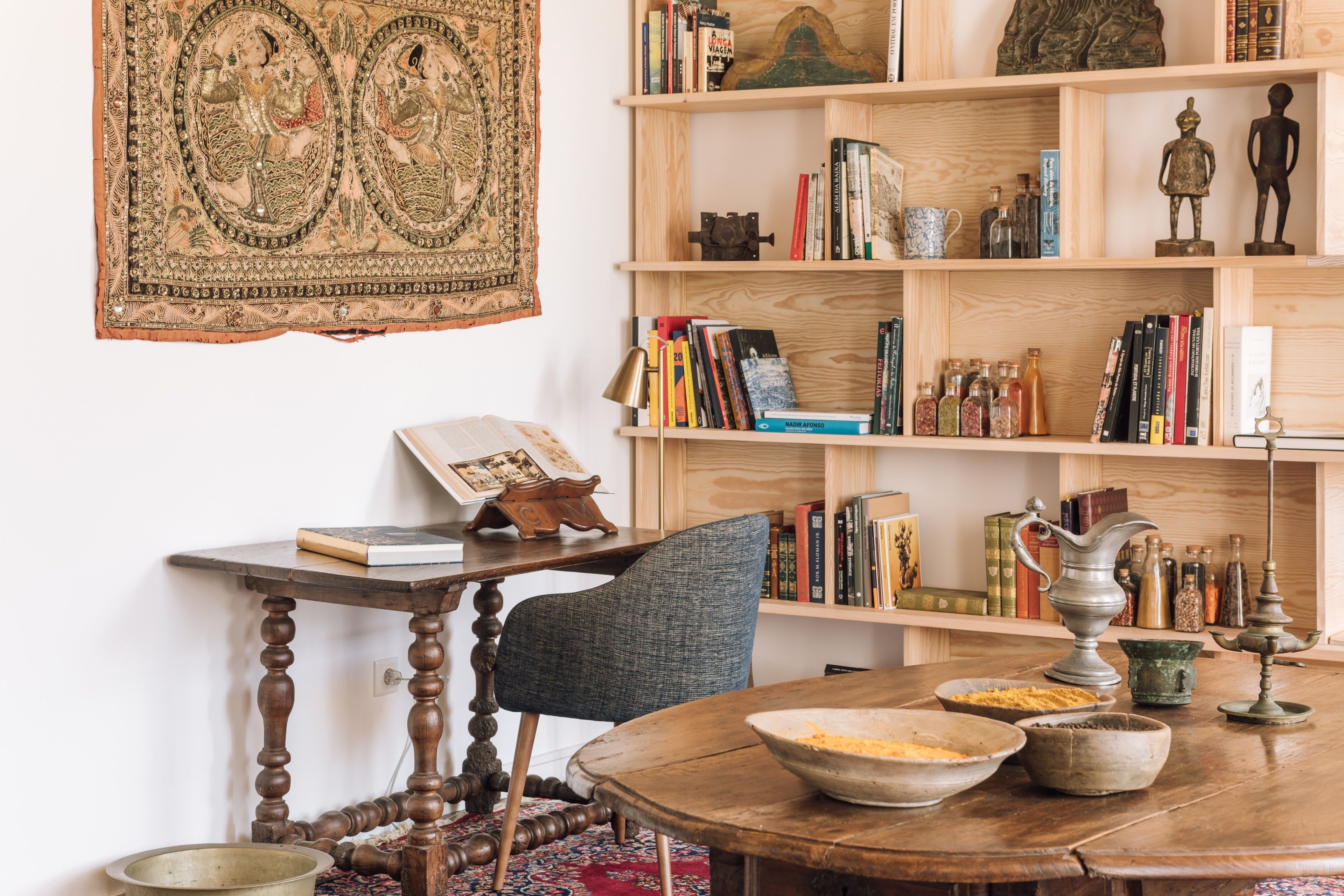 A room with a round wooden table, a bookshelf, and a tapestry.