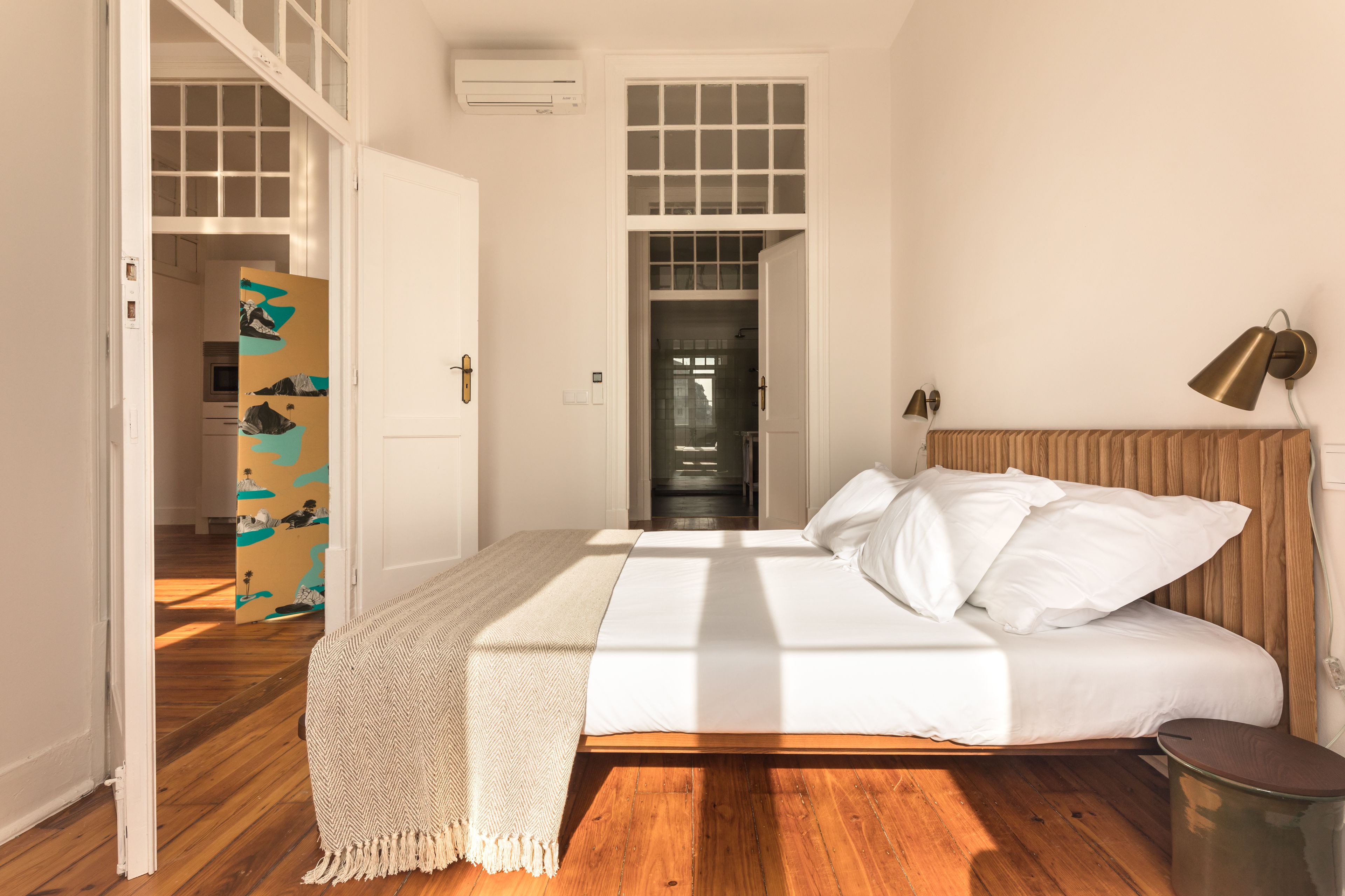 A bedroom with a wooden floor, a bed with white bedding, a throw blanket, and a bedside lamp.