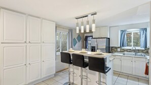Una cocina con armarios blancos, una isla central con taburetes de bar, un refrigerador y una ventana con cortinas.