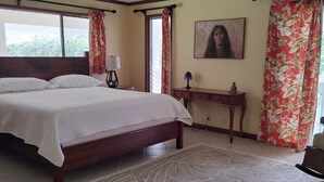 Um quarto com uma cama grande, um ventilador de teto, mesa de cabeceira, abajur, quadro e janela com cortinas.
