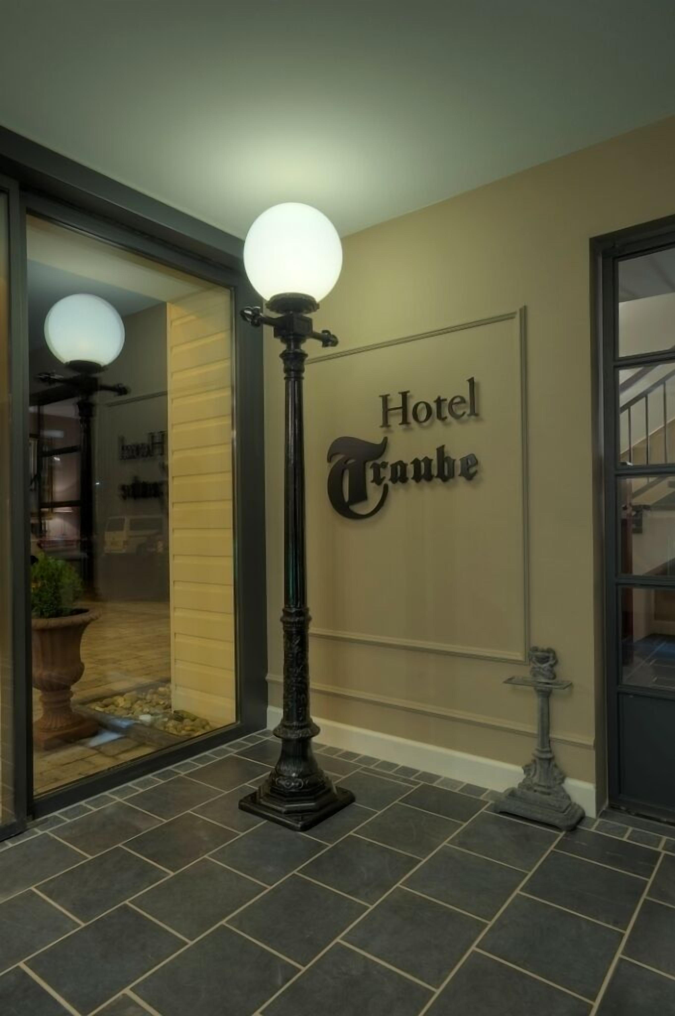 A hotel entrance with a sign reading 'Hotel Traube,' a tall street lamp, and a potted plant.