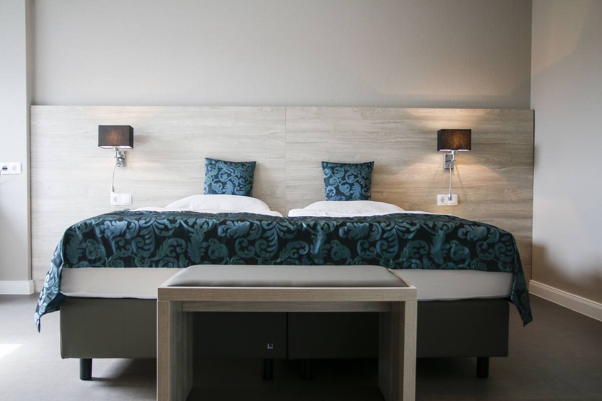 Un lit double avec une couvre-lit à motifs, deux lampes fixées au mur et une table de chevet avec des tiroirs.