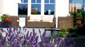 A patio area with a beige sofa, a wicker chair, and a potted plant with purple flowers. - Hotel Zur Traube (Perl)