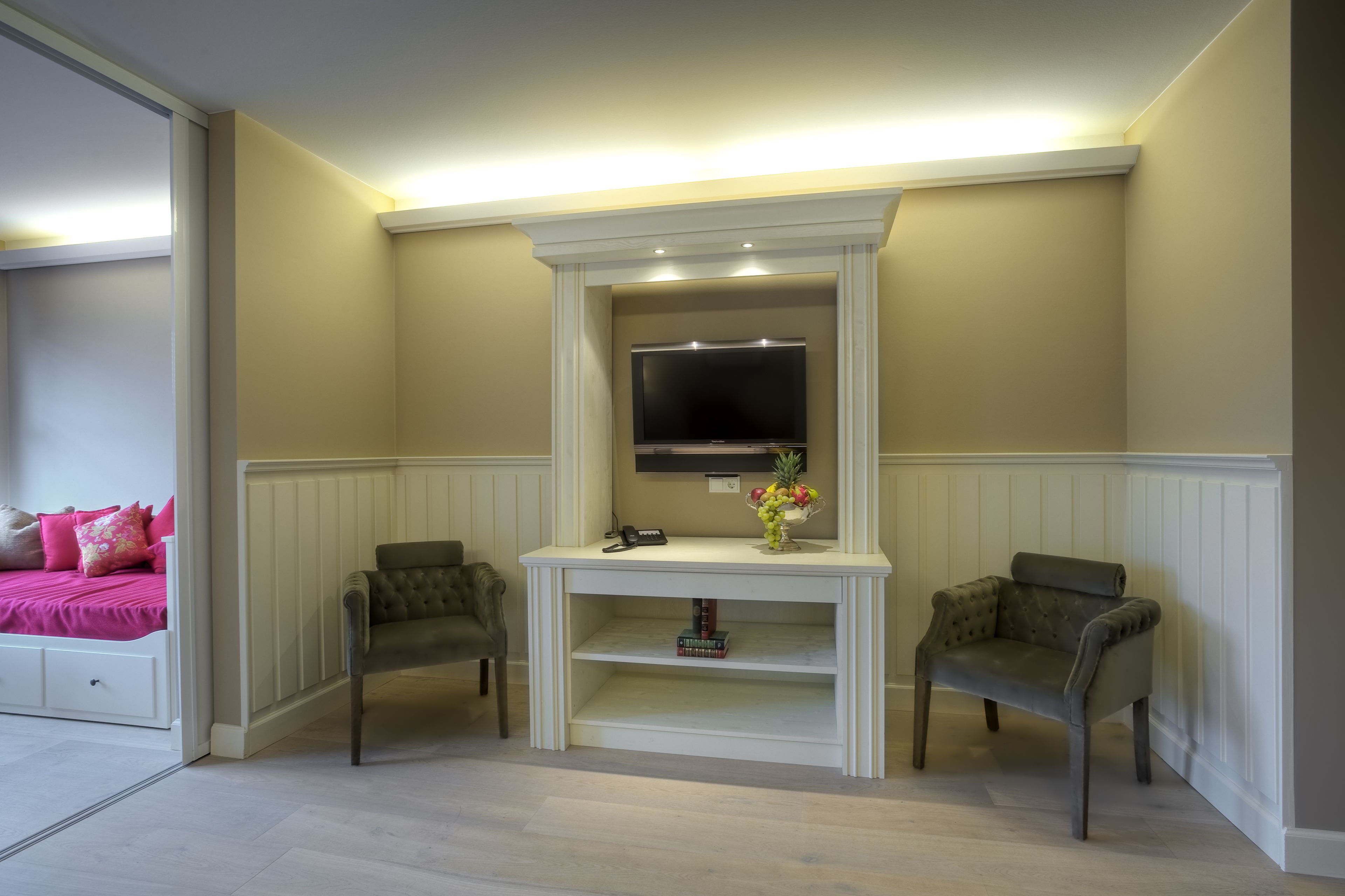 Una habitación con una televisión sobre una mesa consola blanca, dos sillones, una cama rosa y un aparador blanco.