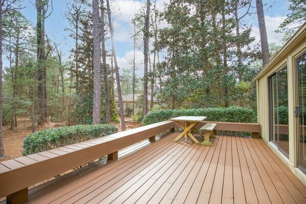 Eine Holzterrasse mit Bänken und einem Tisch, umgeben von Bäumen und einem Gebäude mit großen Fenstern.