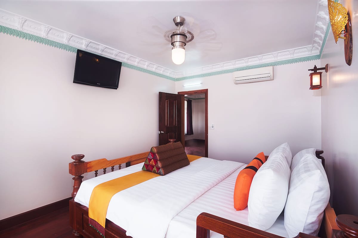 Cama bem arrumada com manta branca e laranja, cabeceira de madeira e duas almofadas. Um ventilador de teto é visível acima da cama, e uma TV de tela plana está fixada na parede.
