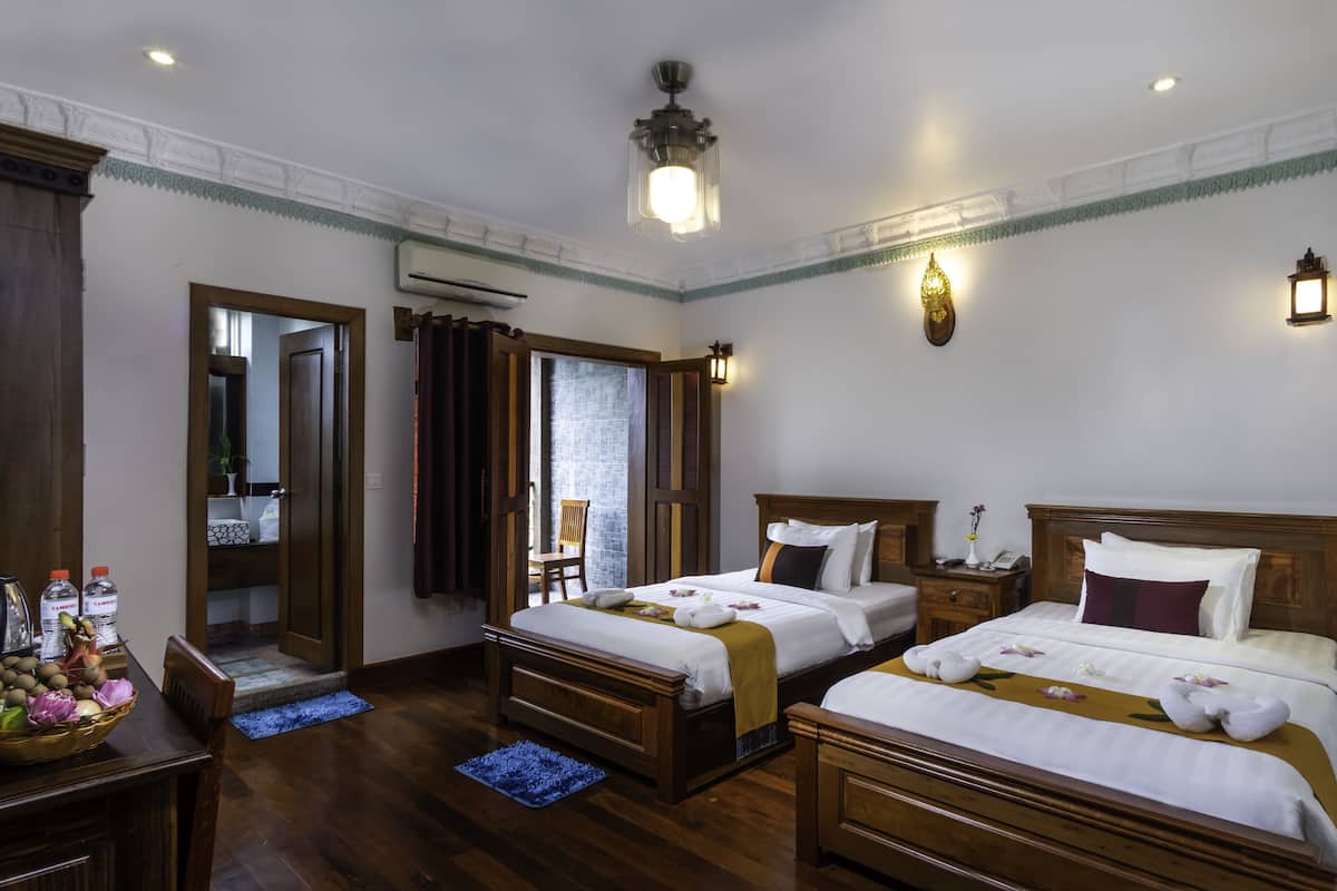 Quarto de hotel com duas camas, móveis de madeira, uma mesa de jantar com frutas e vista para o exterior.