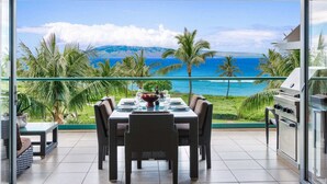 A dining area with a table set for four, overlooking a scenic ocean view with palm trees and mountains. - Maui Resort Rentals: Honua Kai Konea 351, 3BR w/ Direct Oceanfront Views + B.B.Q. on Balcony! (Lahaina)