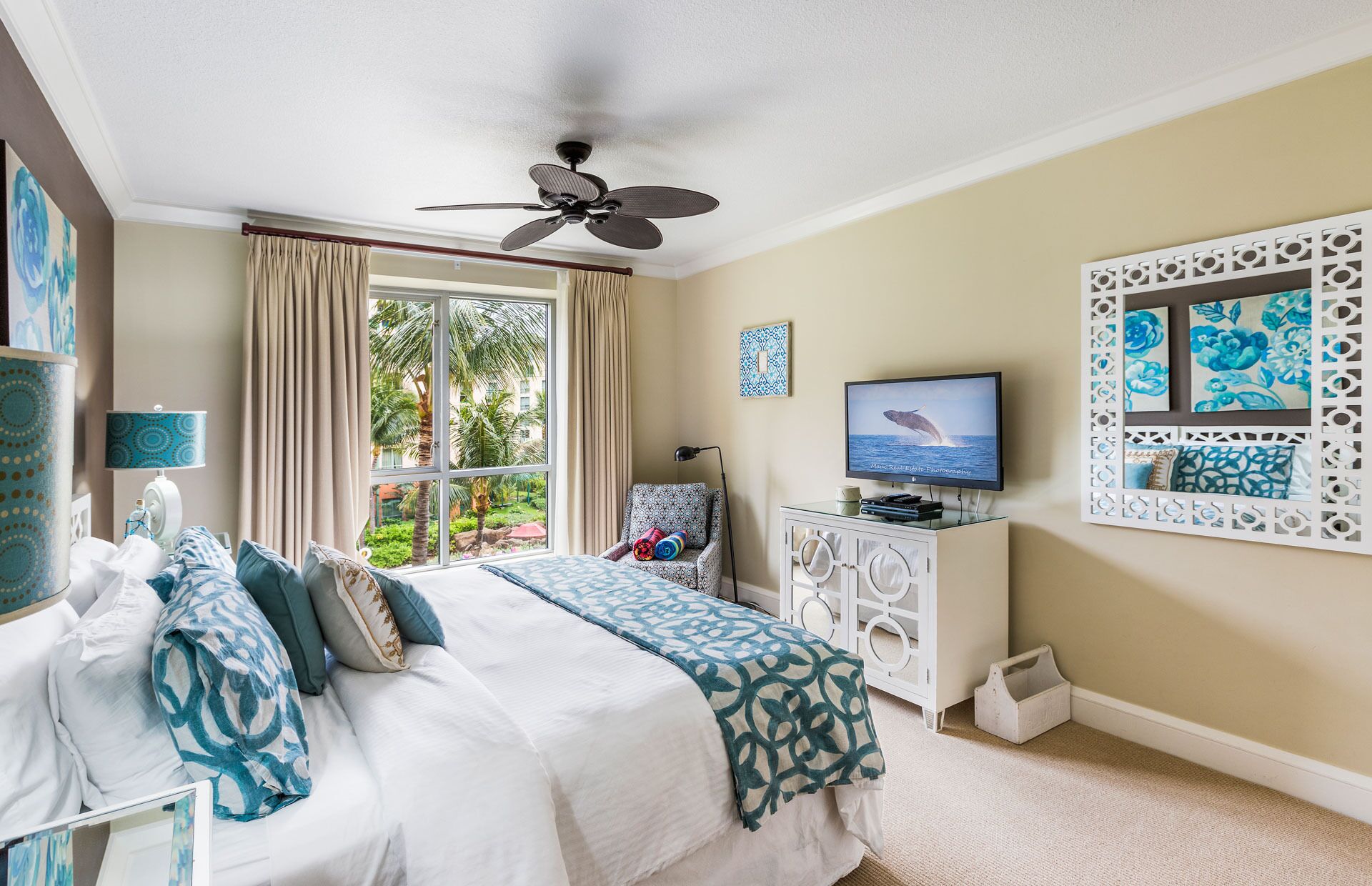 A hotel room with a bed, a television, a ceiling fan, a window with curtains, and a decorative wall panel.
