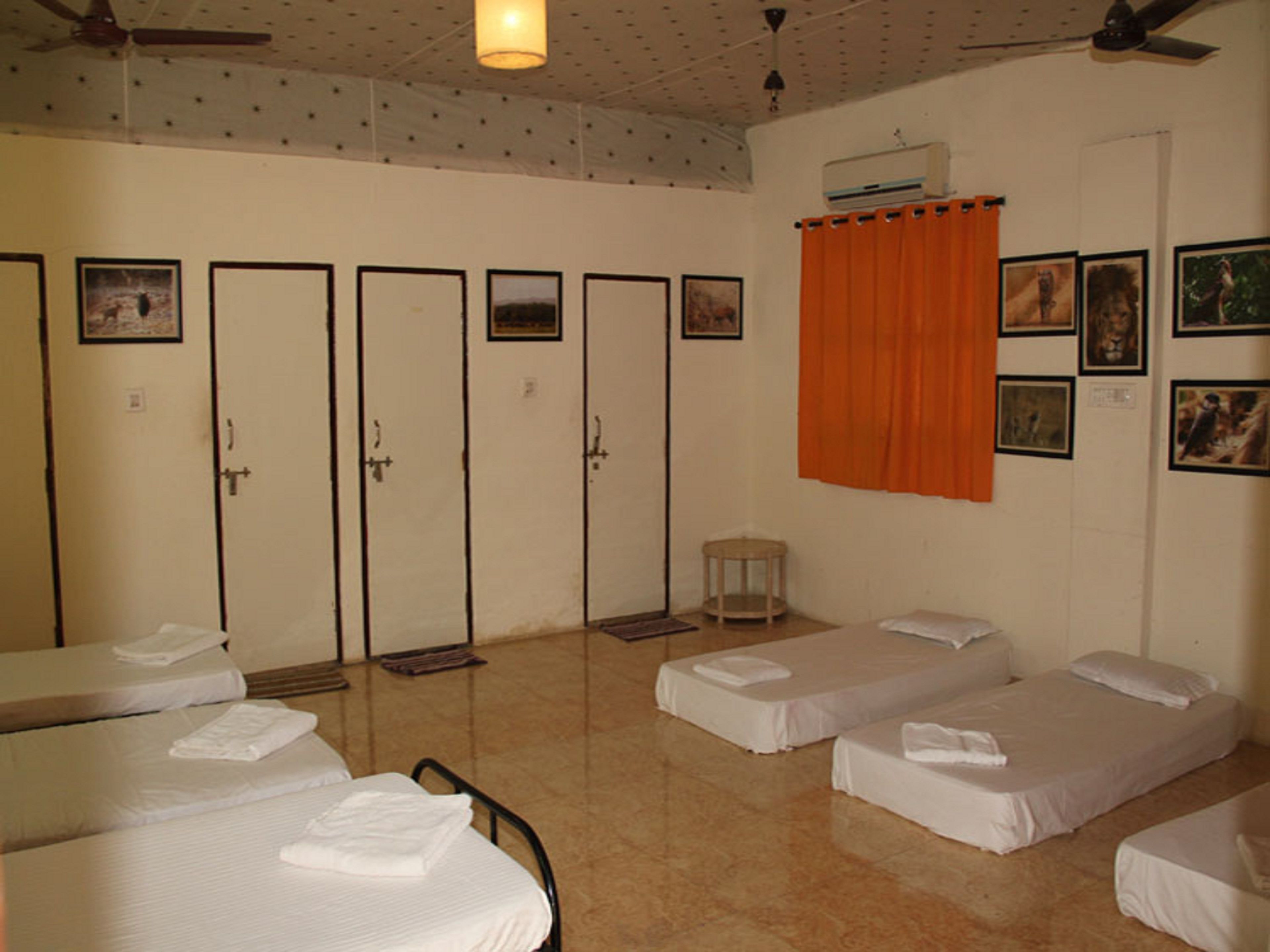 Habitación de hotel con cuatro camas, una mesita, una silla, un ventilador de techo y una ventana con cortina naranja.