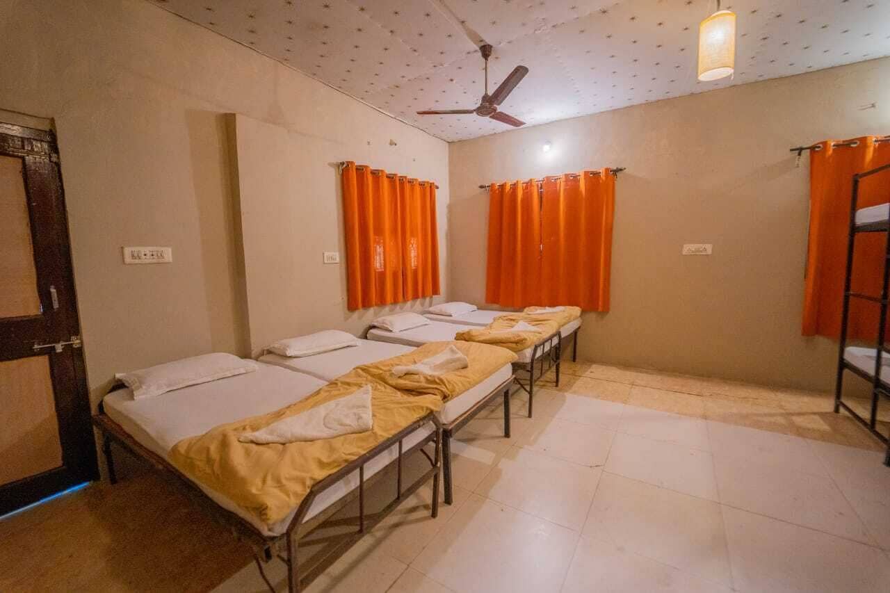 A dormitory room with four single beds, orange curtains, a ceiling fan, and a tiled floor.
