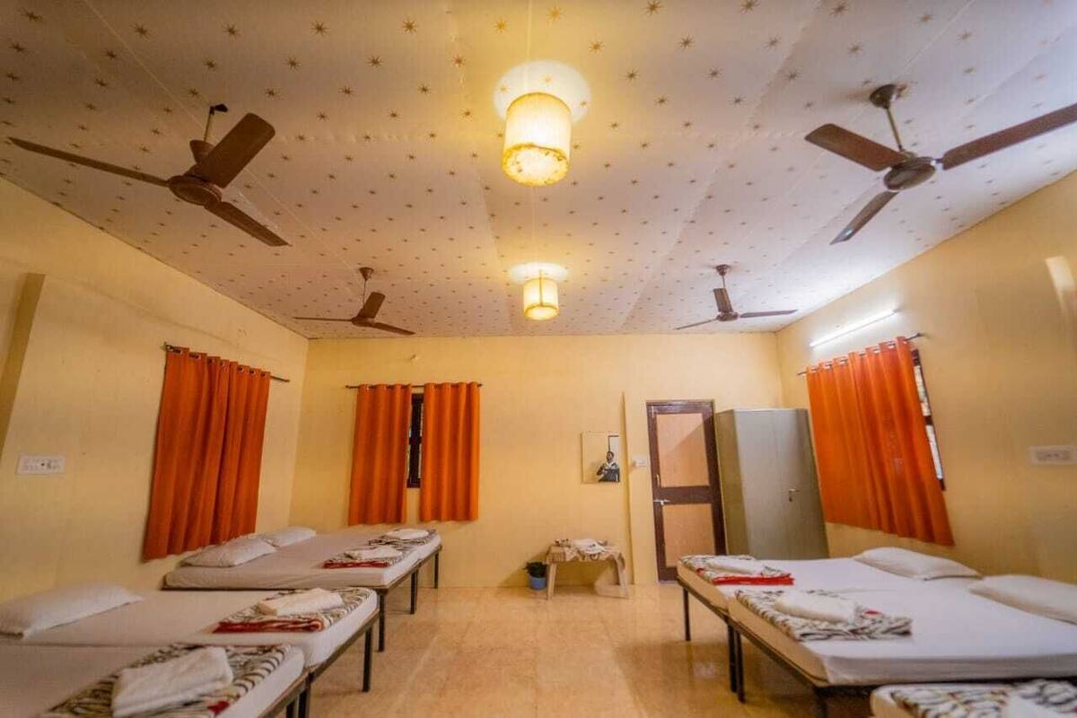 Habitación de hotel con cuatro camas individuales, ventiladores de techo, una mesita con lámpara y cortinas color naranja.