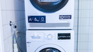 Lavadora y secadora apiladas en un baño con paredes de azulejos blancos y una tubería visible.