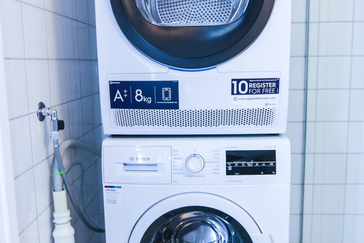 A stacked washer and dryer unit in a bathroom with white tiled walls and a visible pipe.