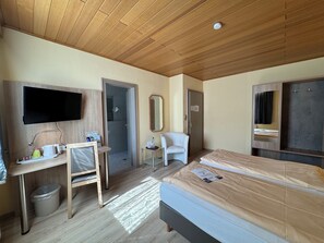 Ein Hotelzimmer mit Holzbalkendecke, einem Bett, einem Schreibtisch mit Stuhl, einem an der Wand befestigten Fernseher und einer Duschkabine.