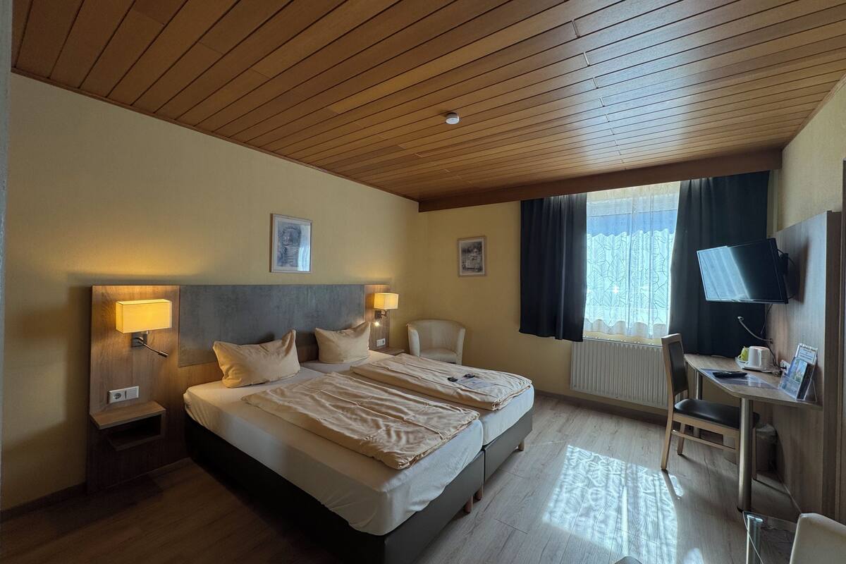 Hotellihuone, jossa on puinen katto, sänky, jossa on kaksi tyynyä, työpöytä tuolilla, televisio ja ikkuna, jossa on verhot.