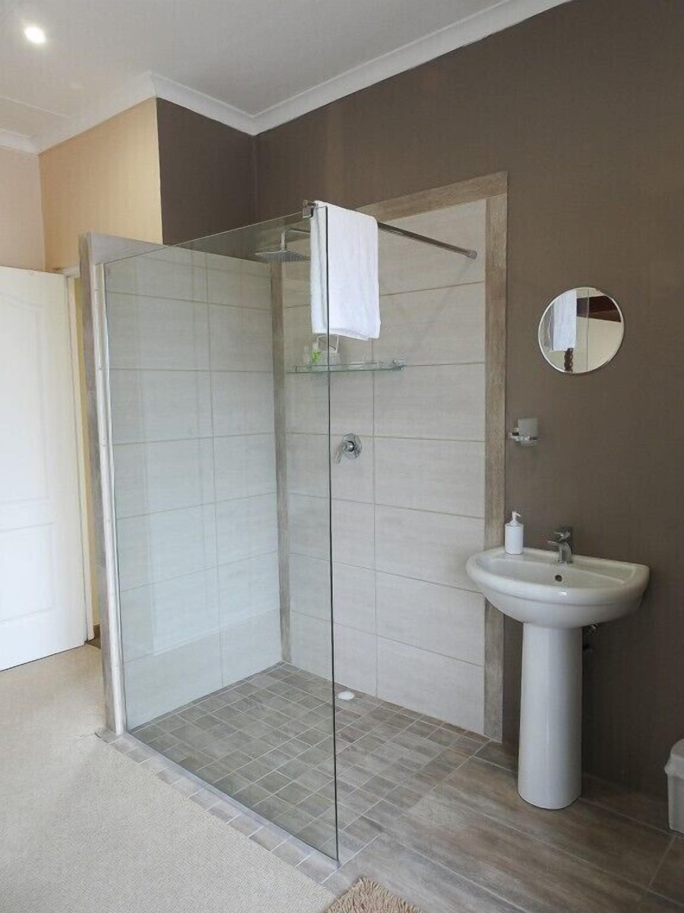 A modern bathroom with a glass shower enclosure, a pedestal sink, and a round mirror.
