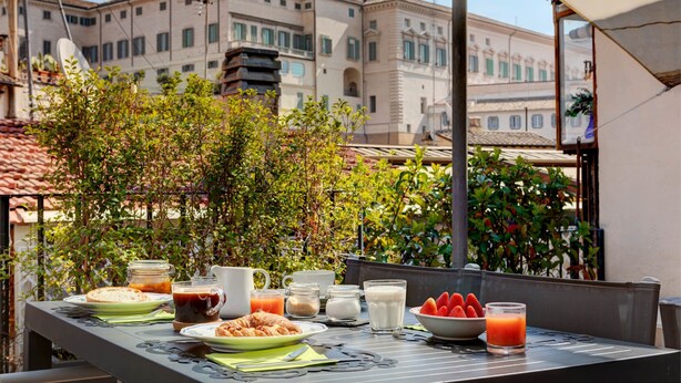 An outdoor dining table with breakfast items, surrounded by greenery and a cityscape in the background. - Casa Modelli (Rome)