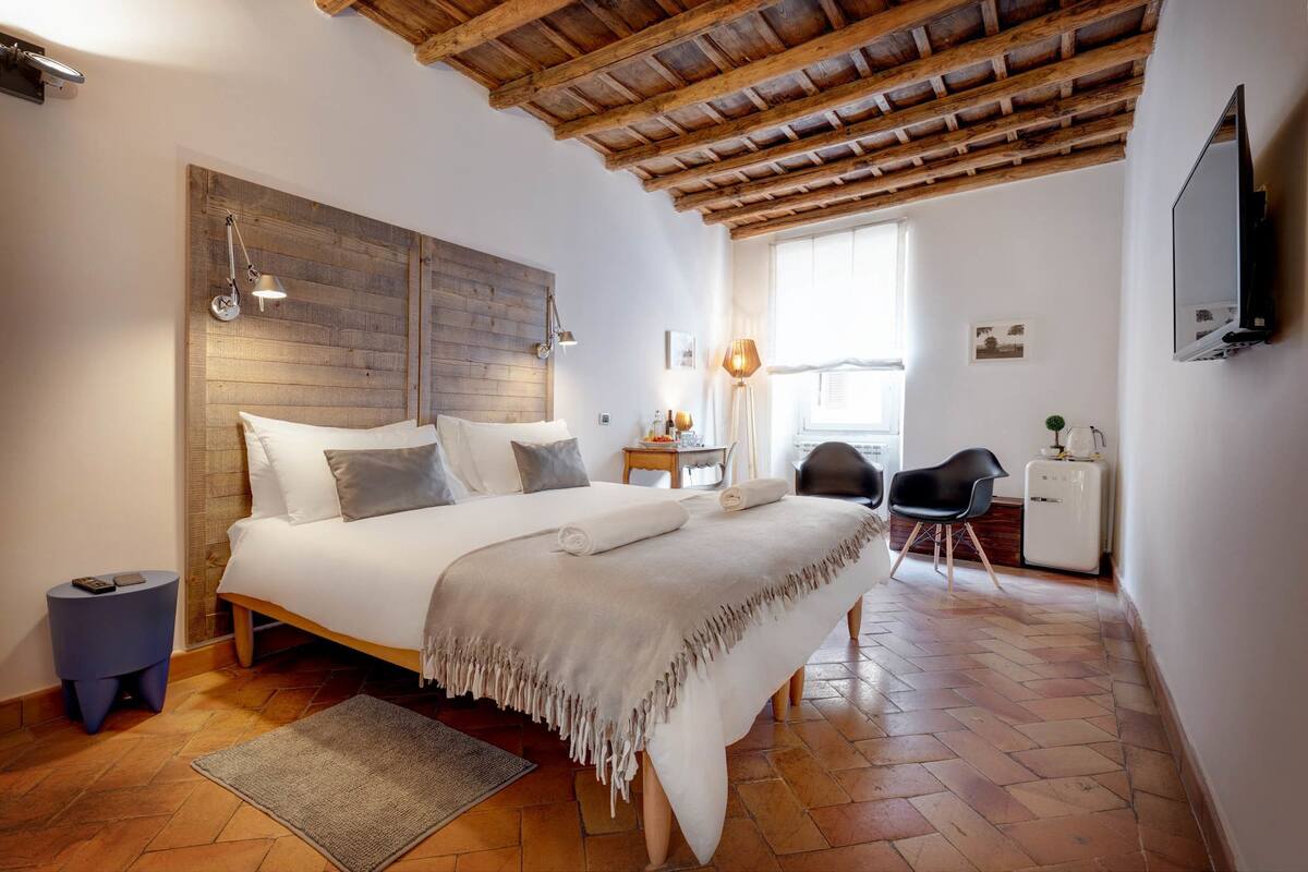 A bedroom with a wooden headboard, a bed with white linens, a desk with a chair, a television, and a window with natural light.