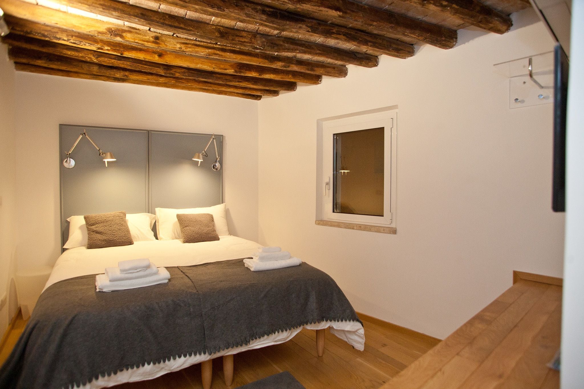 A neatly made bed with a grey blanket and two pillows, a wooden ceiling, a window with a white frame, and a wall-mounted light fixture.