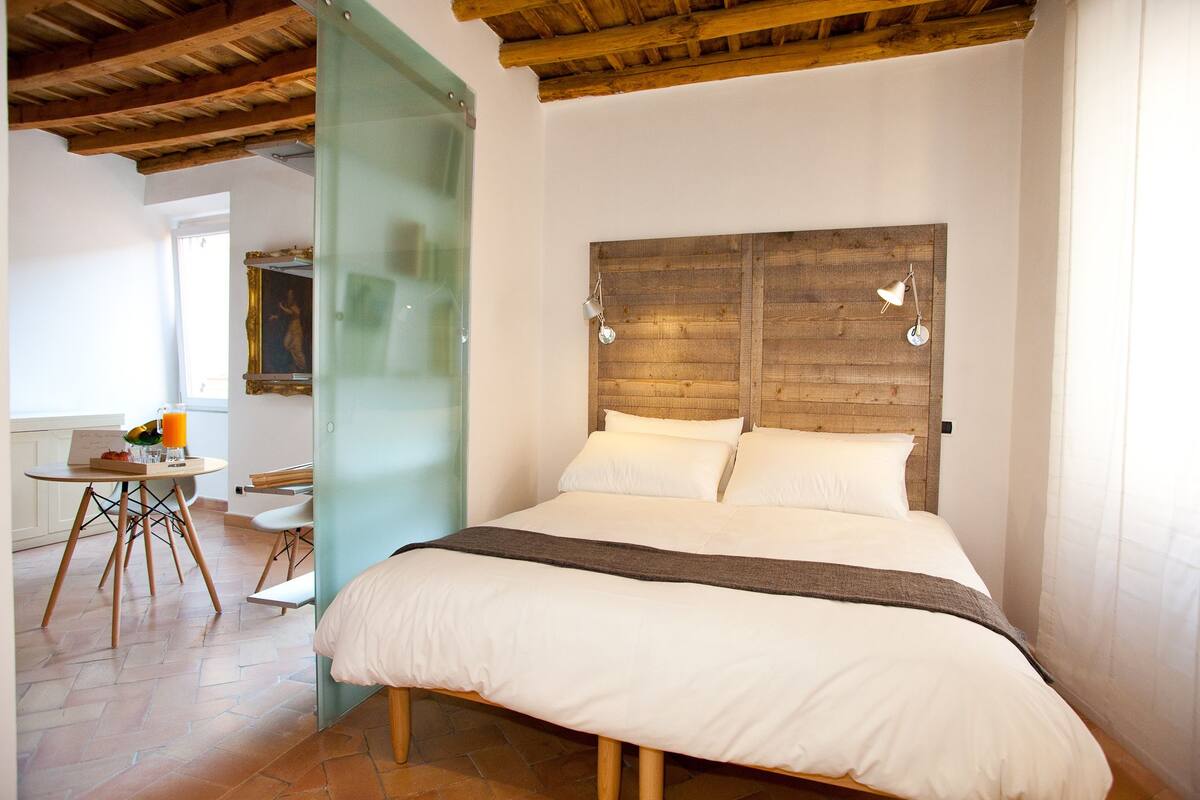 A bedroom with a wooden headboard, a bed with white bedding, a small table with a drink, and a chair.