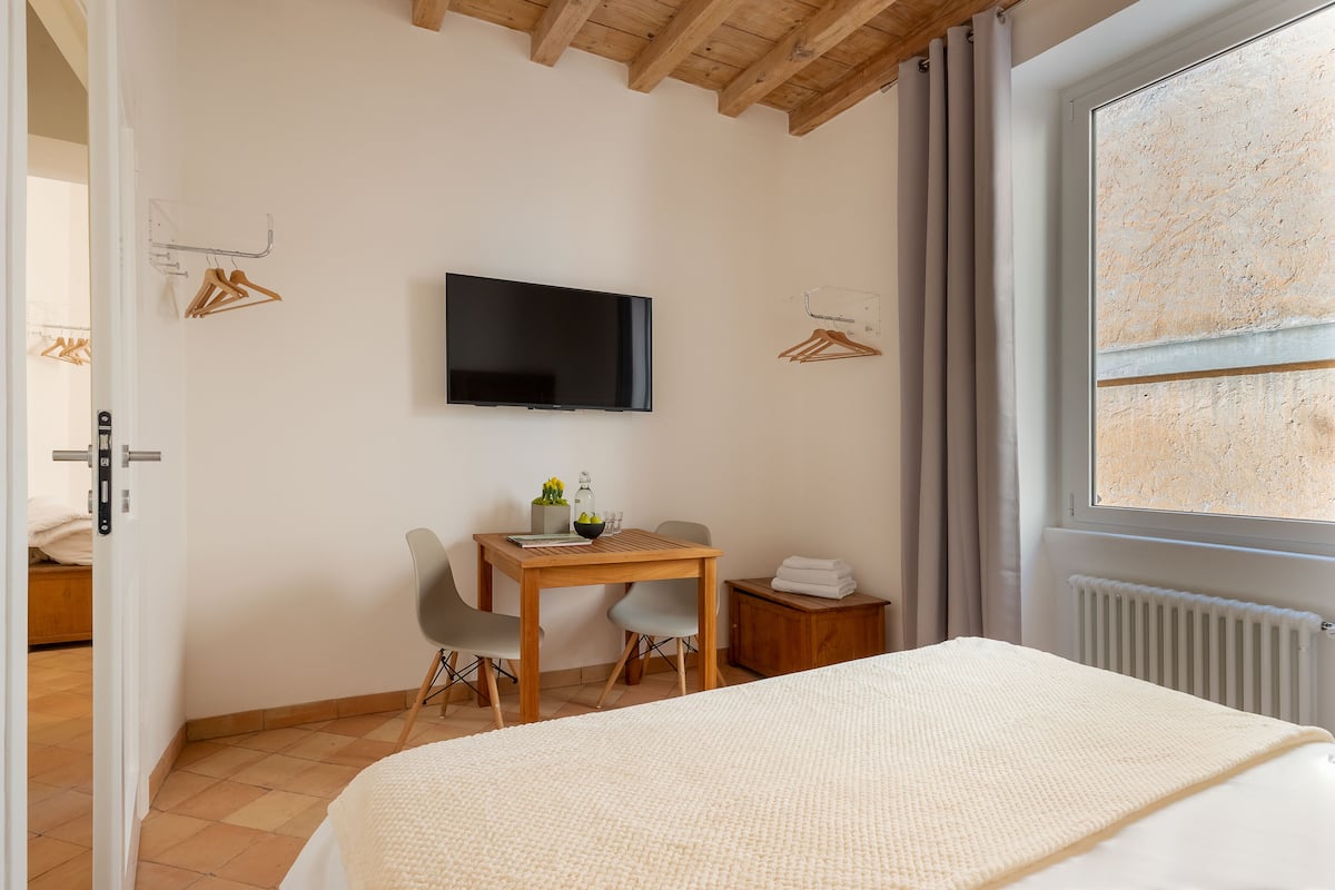 A bedroom with a bed, a desk, a chair, a TV, and a window.