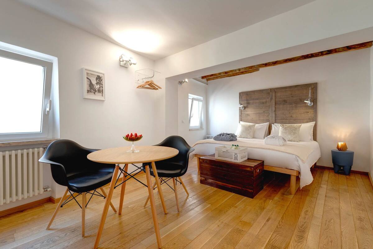 A bedroom with a bed, bedside table, two chairs, a small table with a bowl of fruit, a radiator, and a window.
