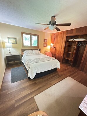 A bedroom with a bed, a nightstand, a lamp, a ceiling fan, a window, and wooden walls. - Cabin/Vacation Home In the Woods on Private Lake Close to Louisville, Ky (Elizabeth)