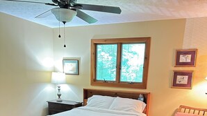 Un dormitorio con cama, una silla, ventilador de techo, ventana y cuadros en la pared.